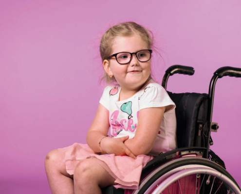 little girl with blonde hair in wheel chair in front of pink background